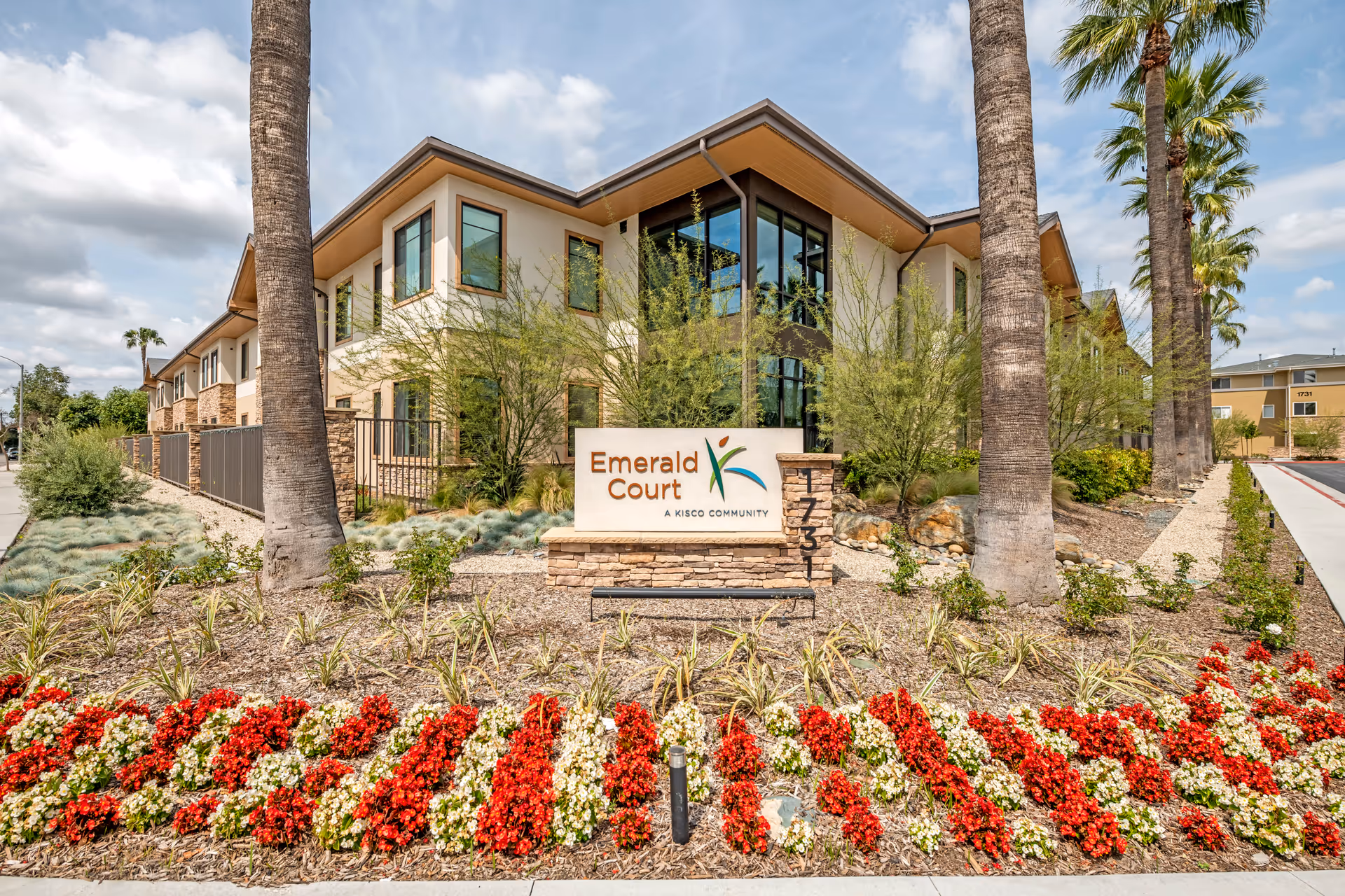 Exterior view of the Emerald Court senior living facility building with a landscaped garden featuring red and white flowers, palm trees, and a sign that reads 'Emerald Court A Kisco Community'. The building has large windows and a modern design under a partly cloudy sky.