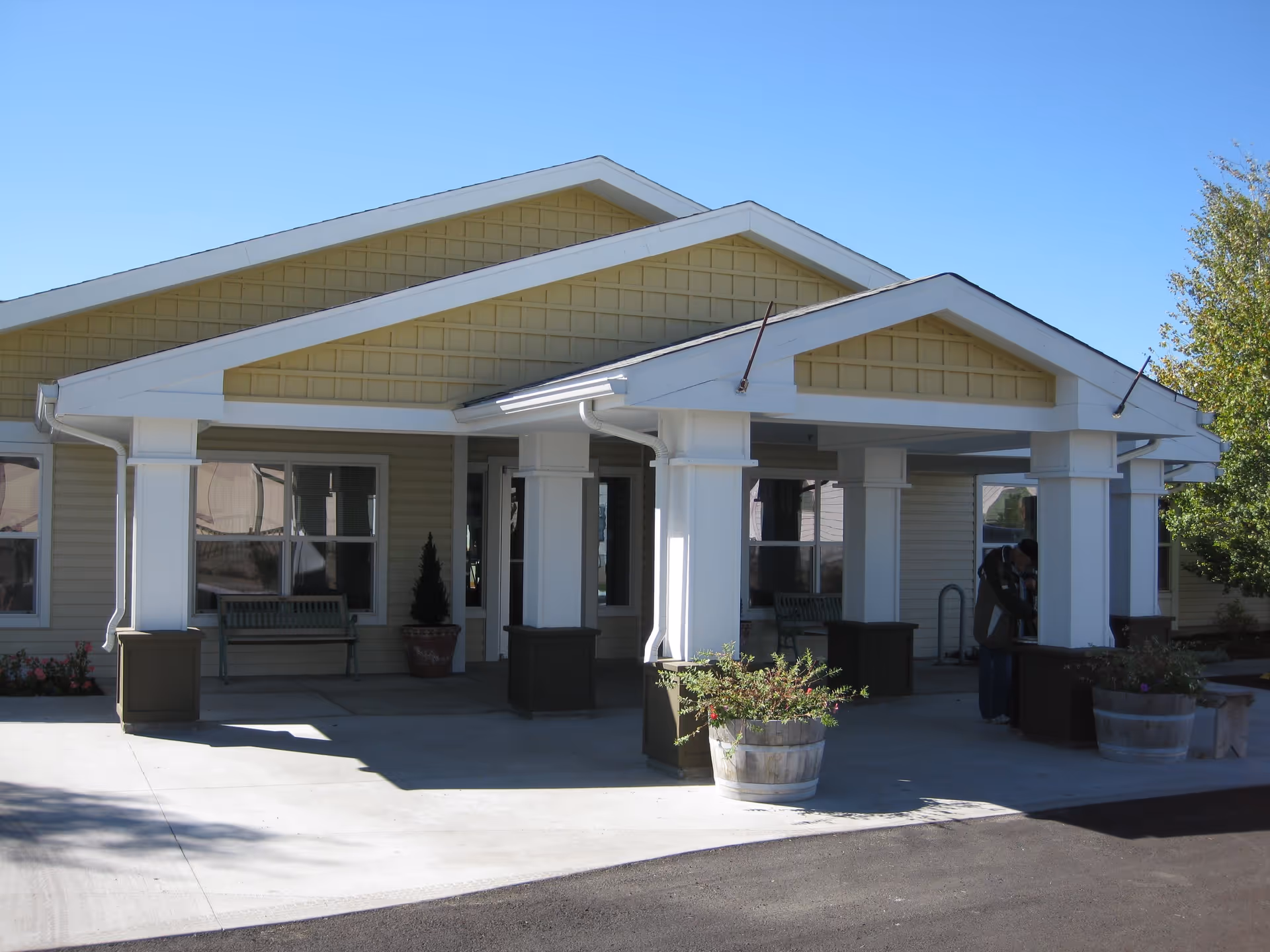 Front exterior view of Prairie House Assisted Living and Memory Care building with a covered entrance supported by white columns, two wooden benches, potted plants, and a person standing near the entrance.