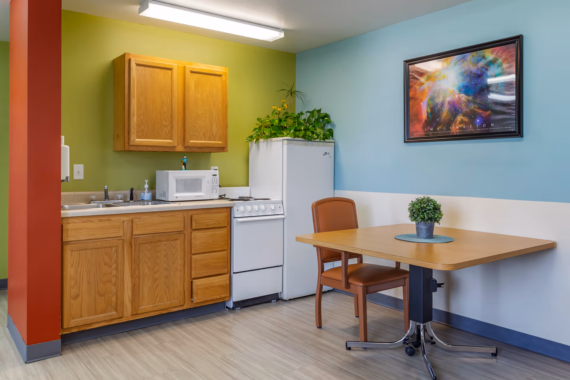 A small kitchen area with wooden cabinets, a white microwave, a white stove, and a white refrigerator topped with green plants. Next to the kitchen is a wooden table with a brown chair and a small potted plant on the table. The walls are painted green and light blue, and there is a colorful framed artwork on the blue wall.