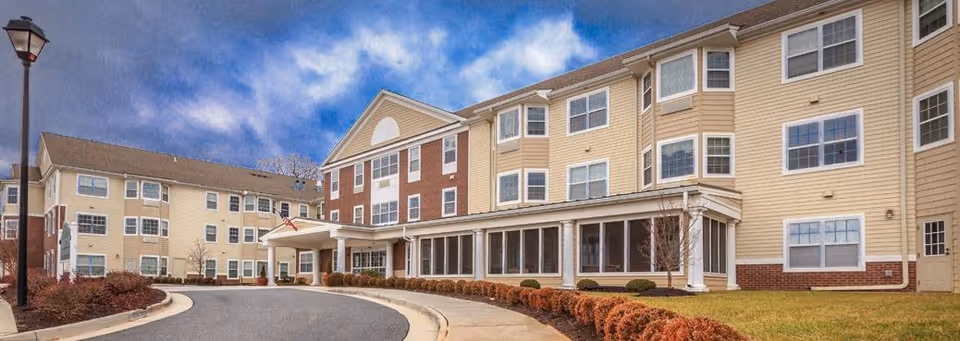 Exterior view of a multi-story senior living facility building with beige siding and red brick accents. The building has many windows and a covered entrance with an American flag. There is a curved driveway and landscaped bushes along the sidewalk under a partly cloudy sky.