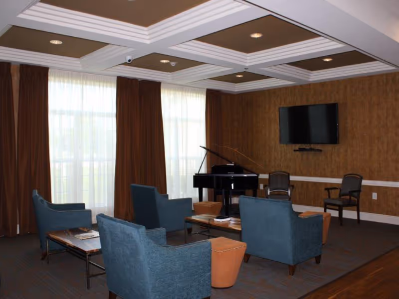 A cozy living room area with four blue armchairs arranged around a coffee table, a black grand piano in the corner, two brown chairs against the wall, a wall-mounted flat screen TV, and large windows with sheer white curtains and brown drapes.