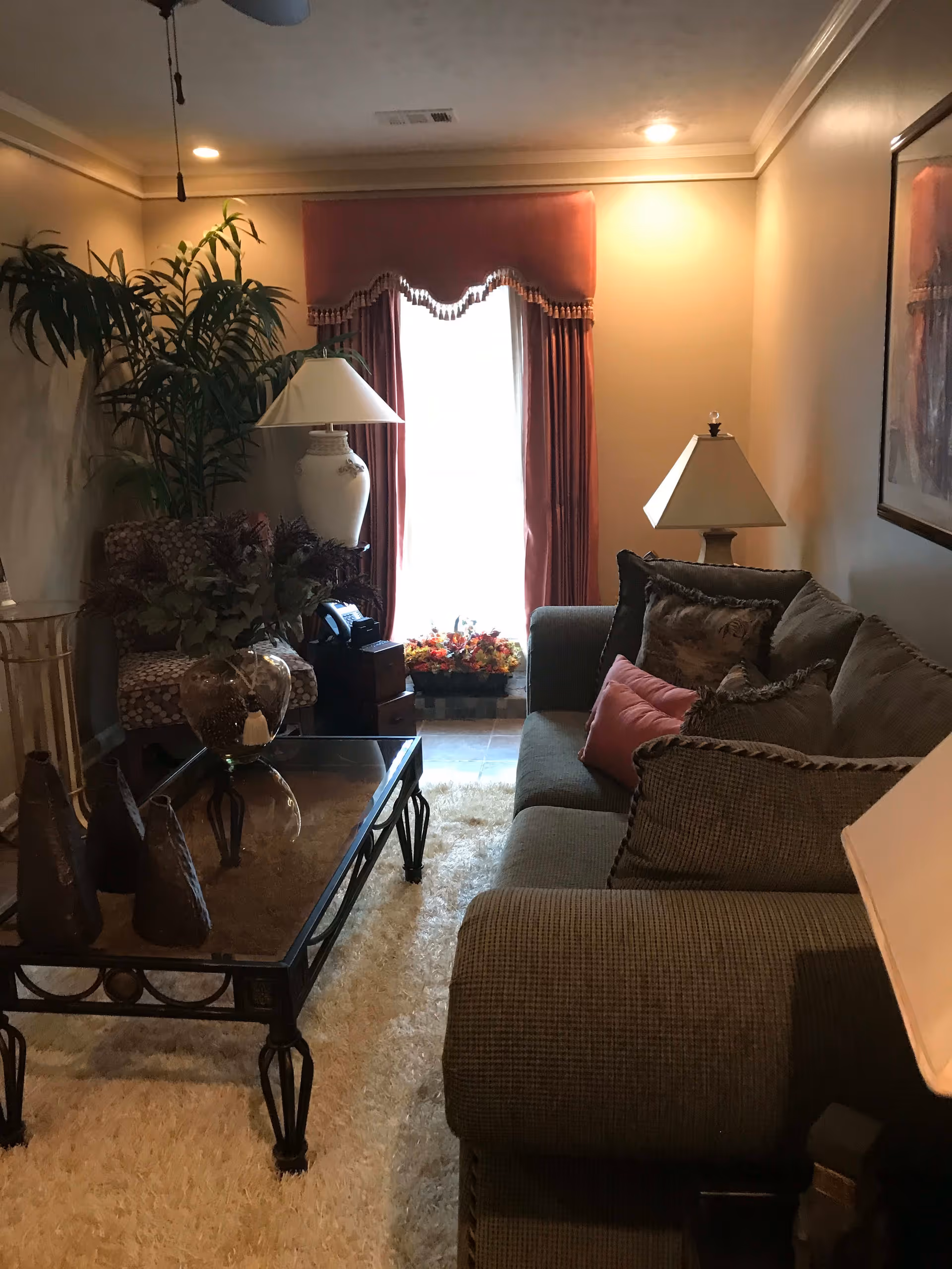 A cozy living room with a large gray sofa adorned with multiple decorative pillows, a glass coffee table with ornamental items, a patterned armchair, two table lamps, a tall potted plant, and a window with red curtains letting in natural light.