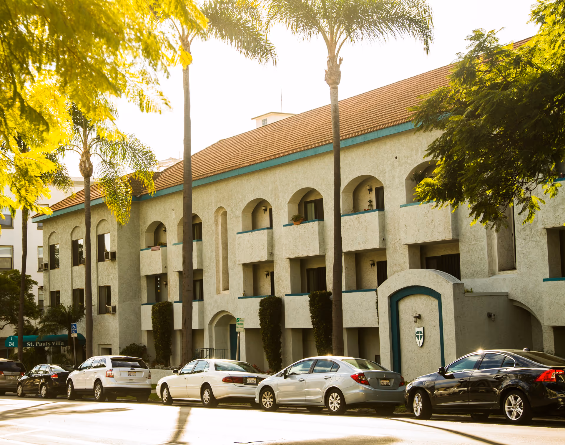 Exterior view of a multi-story residential building with arched balconies and a tiled roof, lined with palm trees and parked cars along the street in front.