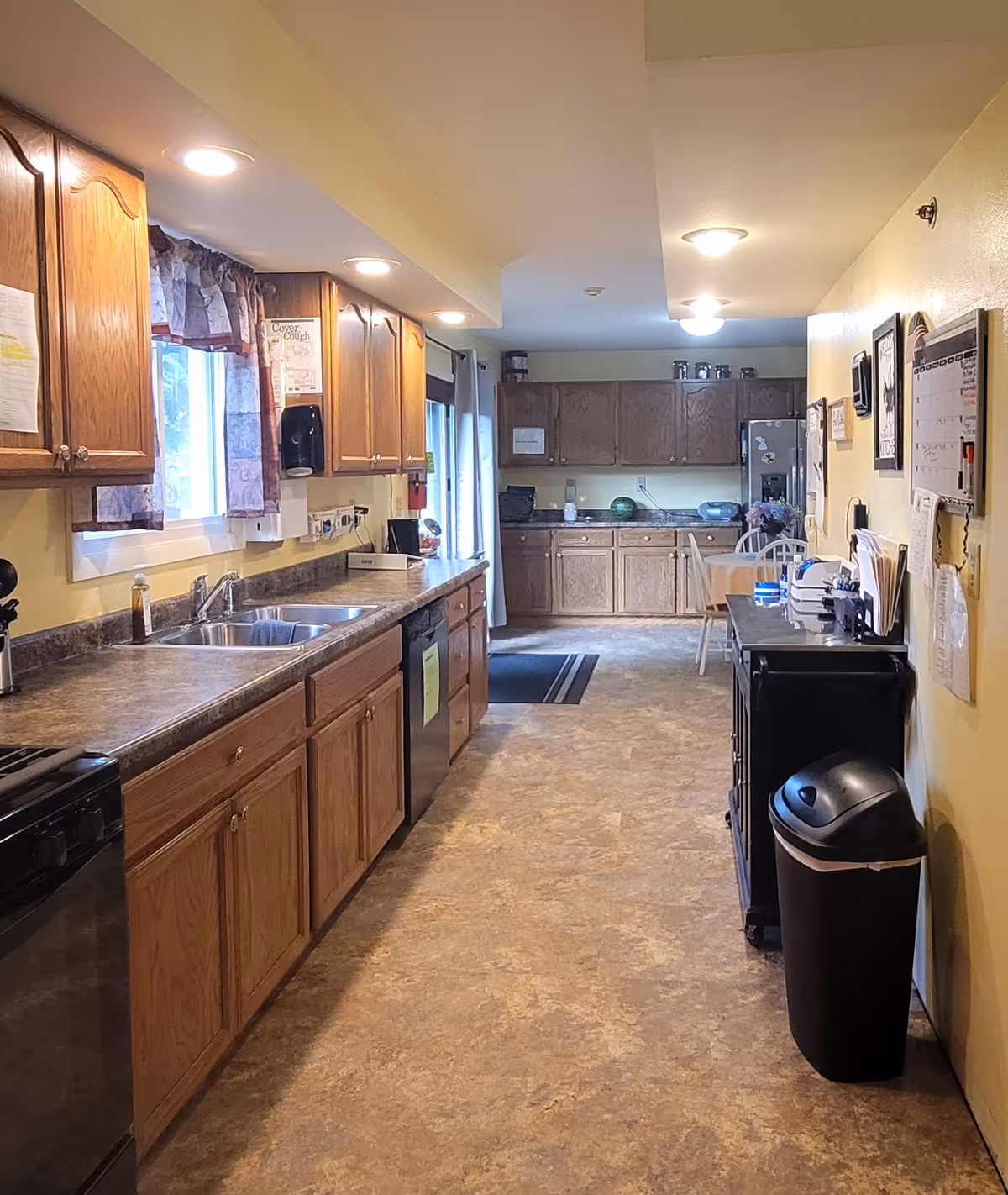 A long kitchen area in an assisted living facility with wooden cabinets, a double sink, countertop appliances, and a trash bin. The kitchen has a window with curtains, overhead lighting, and a small dining table with chairs in the background.