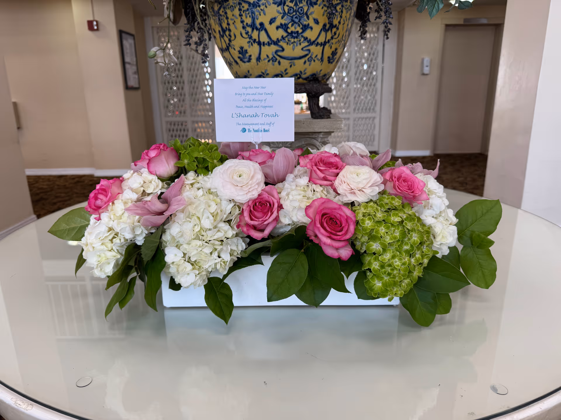 A floral arrangement featuring pink roses, white hydrangeas, green hydrangeas, and assorted greenery placed on a round glass table in an indoor setting with beige walls and carpeted floors. A decorative blue and yellow vase is visible in the background along with a card that reads a greeting message.