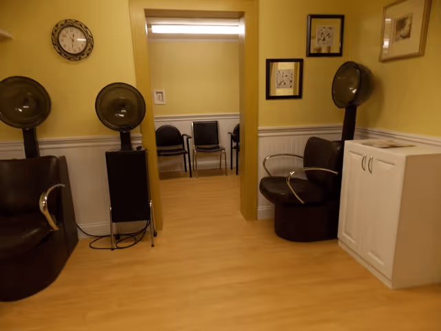 Small salon-style room with hooded hair dryers, chairs, and a doorway opening to a waiting area.