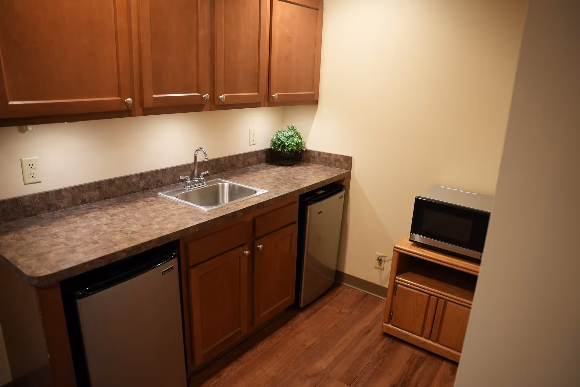 Small kitchenette area with wooden cabinets, a countertop with a sink, a small refrigerator, a mini fridge, and a microwave on a wooden stand. The floor is wooden and the walls are light-colored.