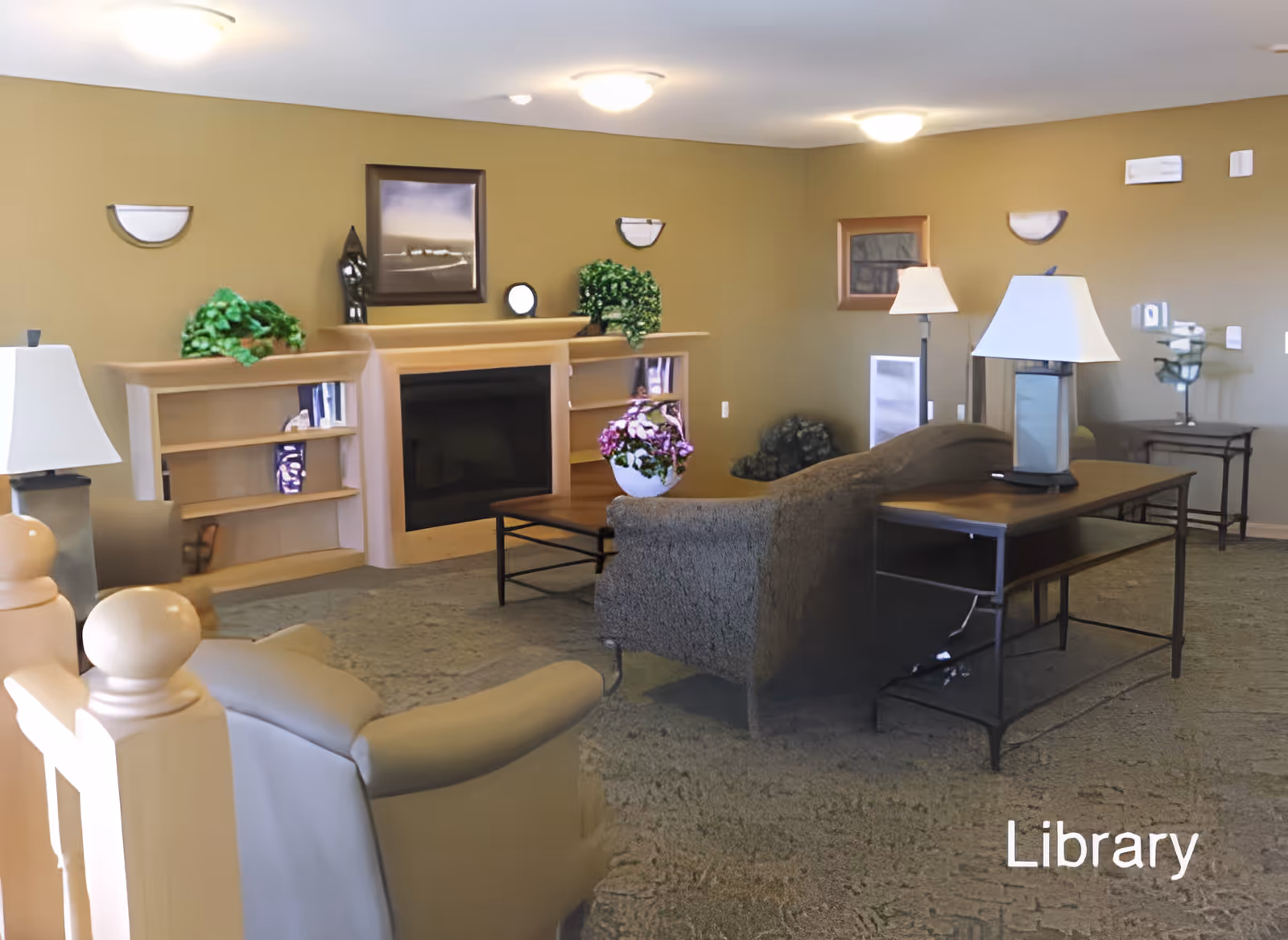 A cozy library room with beige walls and carpeted floor. The room features a fireplace with built-in shelves on either side, decorated with green plants and a framed picture above. There are comfortable armchairs and a sofa arranged around a wooden coffee table with a flower pot. Two table lamps and a floor lamp provide lighting, and there is a small side table with a decorative item in the corner.