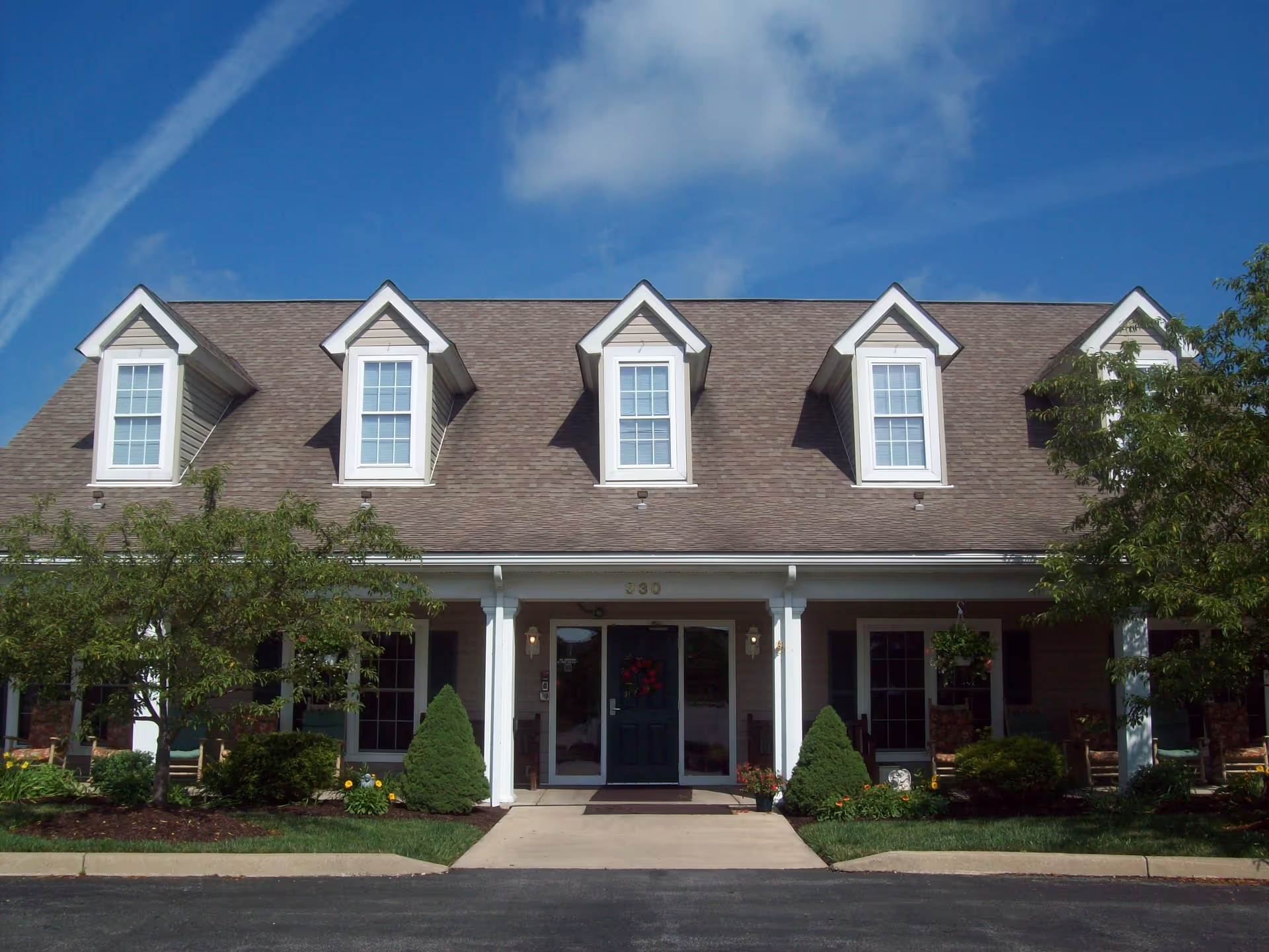 Front exterior of a two-story residential building with dormer windows, a central entrance, and landscaped shrubs under a blue sky.