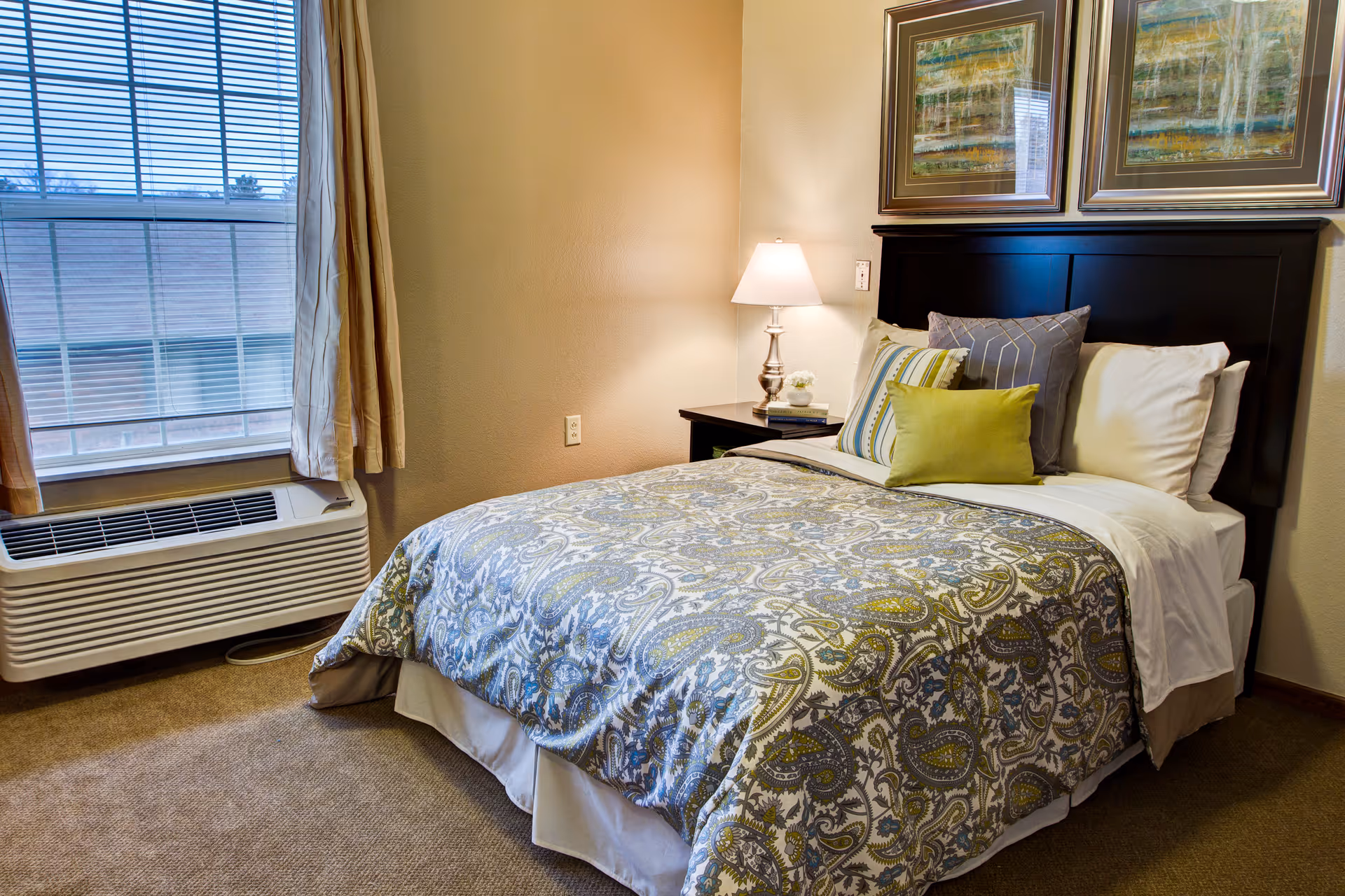 A neatly made bed with a patterned bedspread and multiple pillows in a cozy bedroom. There is a nightstand with a lamp and some books next to the bed. Two framed abstract paintings hang on the wall above the headboard. A window with blinds and curtains is on the left side, and an air conditioning unit is below the window.