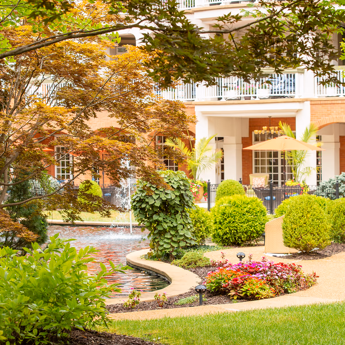 Landscaped courtyard with a curved pond and fountain, trimmed shrubs and flower beds, and a brick building with balconies and an outdoor umbrella in the background.