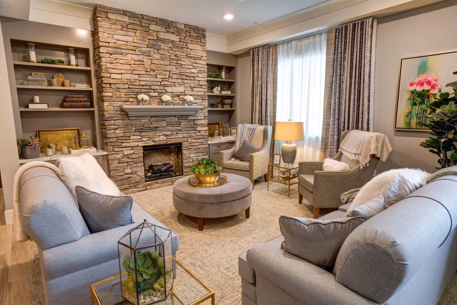 A cozy living room with two light gray sofas, two armchairs, a round ottoman, and a stone fireplace. The room features built-in shelves with decorative items, a window with curtains, a side table with a lamp, and a colorful floral painting on the wall.