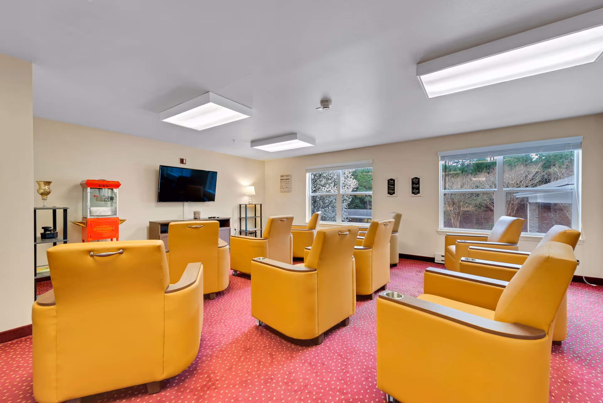 A bright TV lounge with yellow armchairs arranged facing a wall-mounted television, windows, and a popcorn machine.