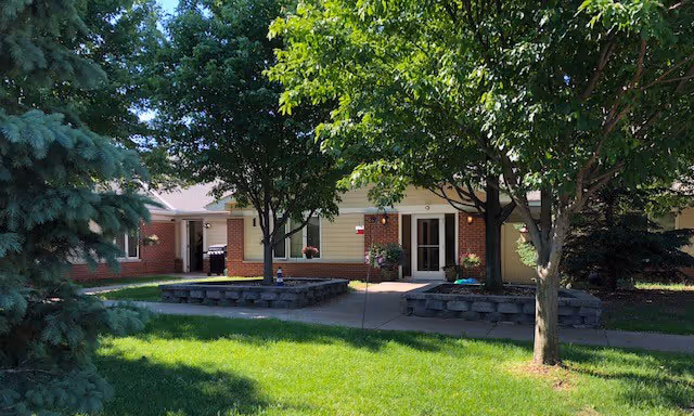 Exterior view of Southern Oaks Place showing a single-story brick and siding building partially shaded by large leafy trees, with a well-maintained green lawn and a paved walkway leading to the entrance.
