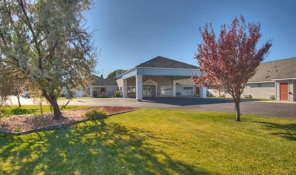 Exterior view of Dorian Place Assisted Living facility showing a single-story building with a covered entrance, surrounded by green grass, trees, and a clear blue sky.