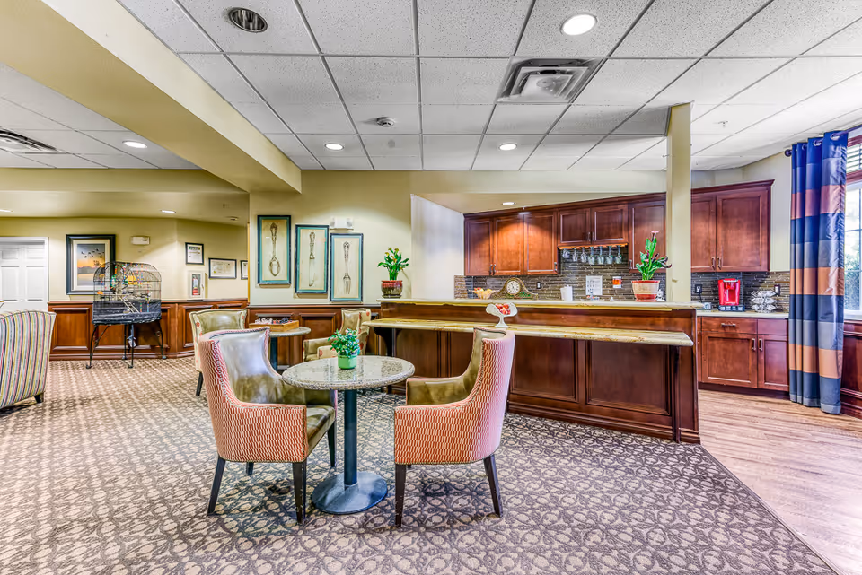 A cozy senior living common area featuring a small round table with four upholstered chairs, a wooden counter with bar stools, and a kitchen area with wooden cabinets and a coffee maker. The room has patterned carpet, framed artwork on the walls, potted plants, and large windows with striped curtains allowing natural light.