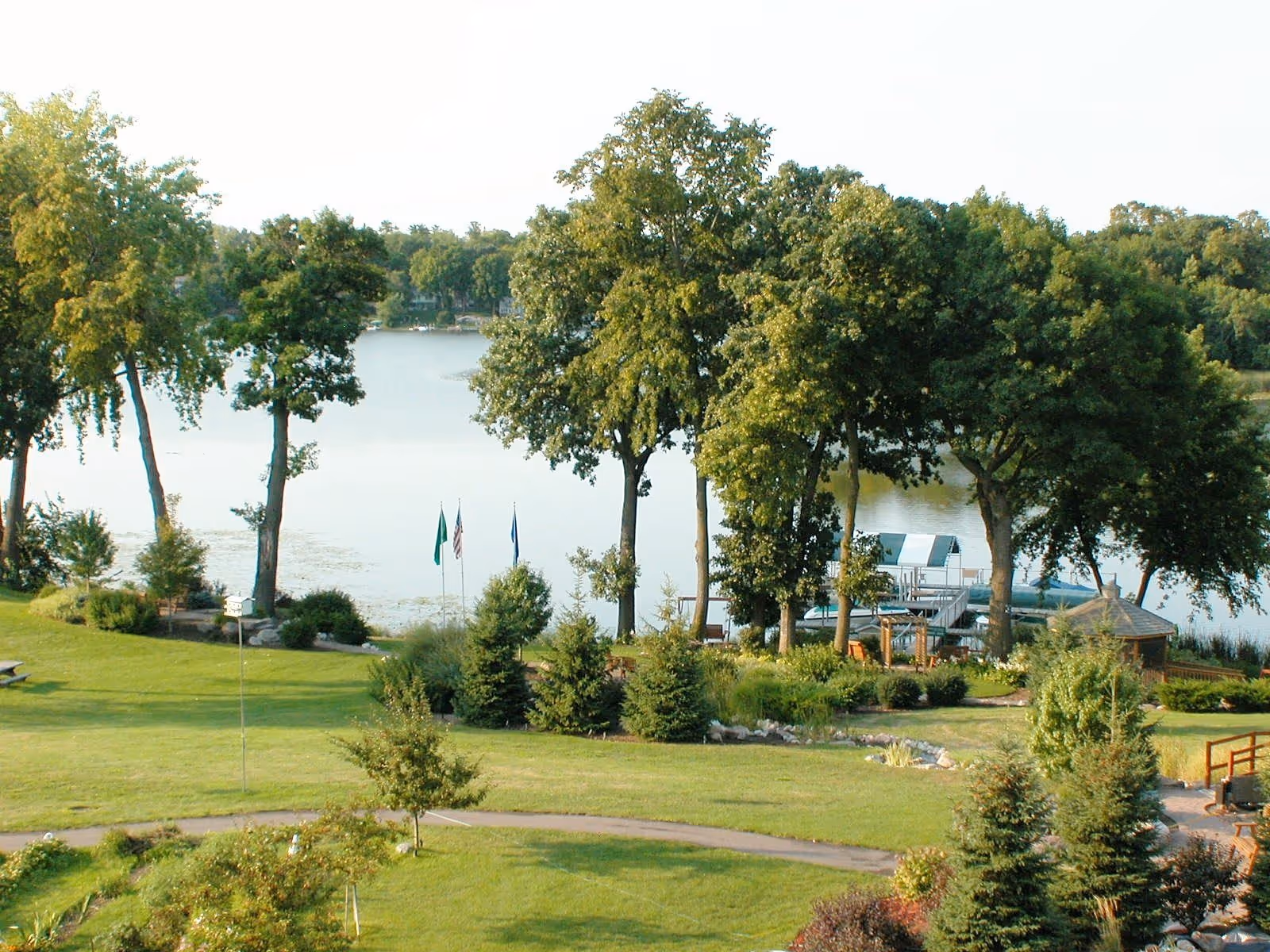 A scenic outdoor view of a lakeside area with green grass, trees, and a dock extending into the water. There are flags near the shore and a small gazebo visible among the trees.