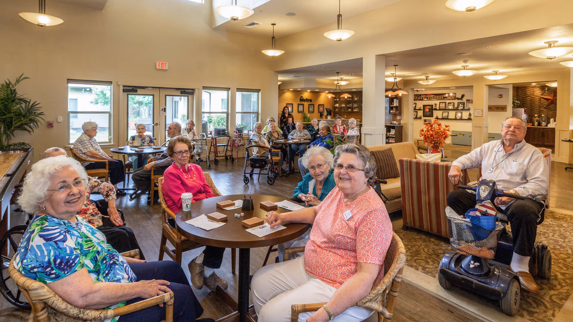 A group of elderly people sitting around tables and in chairs in a well-lit common area of a senior living facility, some playing games and others socializing. The room has large windows, warm lighting, and comfortable seating. A man on a mobility scooter is also present.