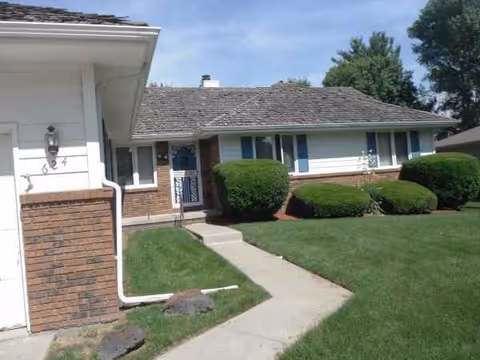 Front exterior of a single-story senior living home with a walkway and trimmed shrubs.