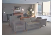 A cozy living room area with a patterned sofa and two matching armchairs arranged around a wooden coffee table. There is a fireplace with a framed picture above it, a floor lamp, and a potted plant near a large window letting in natural light.