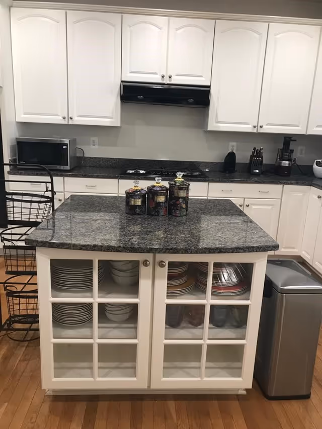 A clean kitchen with a granite island, white cabinets, a stovetop and microwave, and glass-front storage showing dishes.