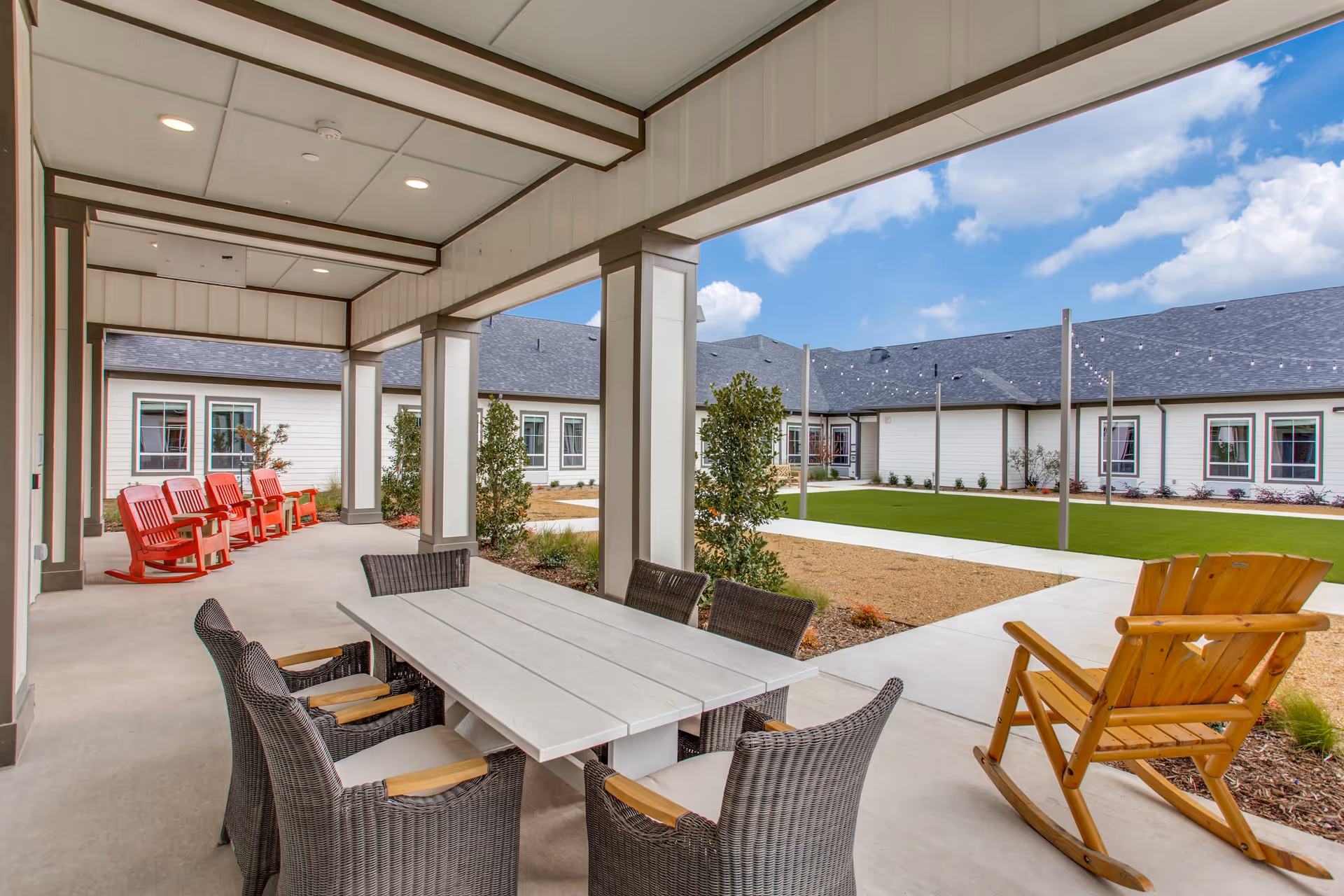 Covered outdoor patio area with a white rectangular table surrounded by six wicker chairs with cushions. To the left, there are four red rocking chairs lined up against the wall. On the right side, there is a wooden rocking chair facing a courtyard with green grass, small bushes, and a building with white siding and multiple windows under a blue sky with some clouds.