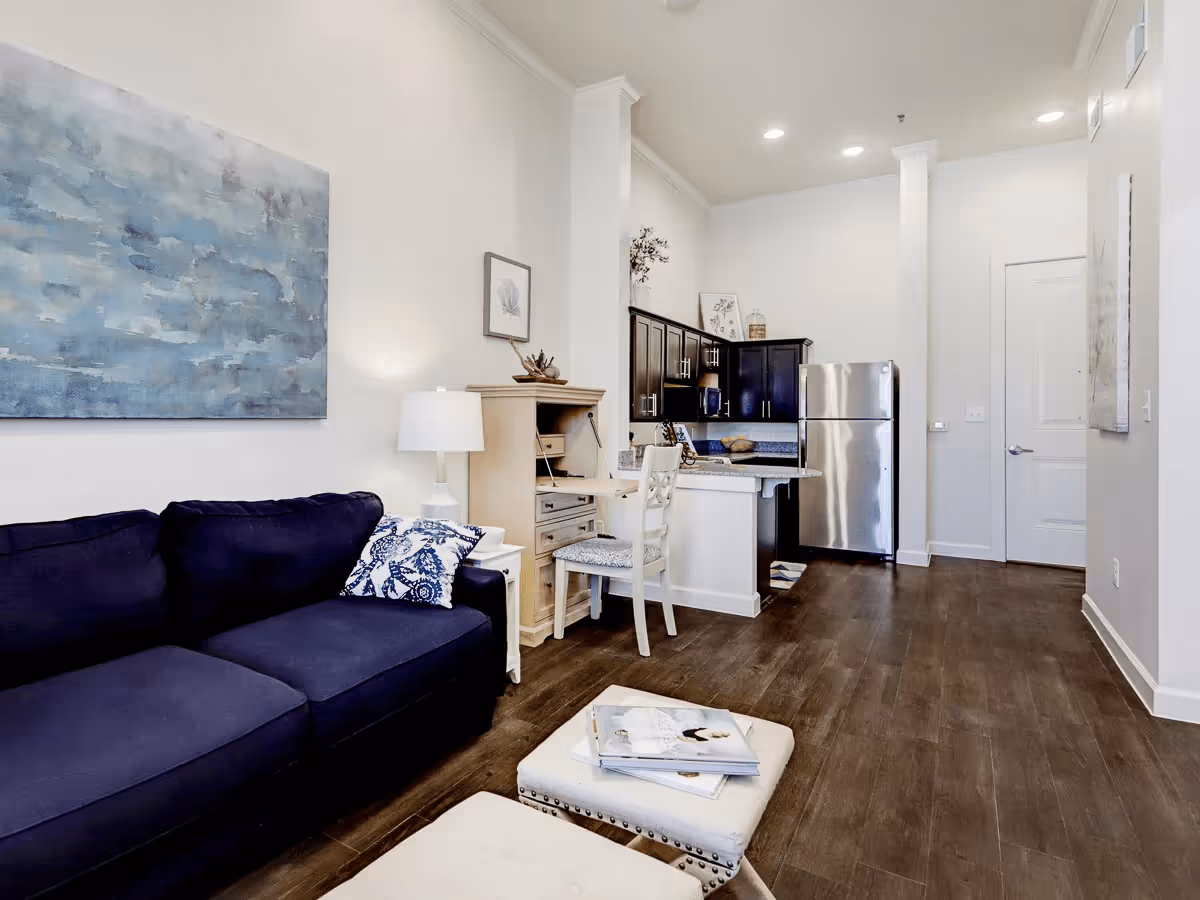 Open-concept living area with a navy sofa, ottoman, small desk and a kitchenette with stainless steel refrigerator.