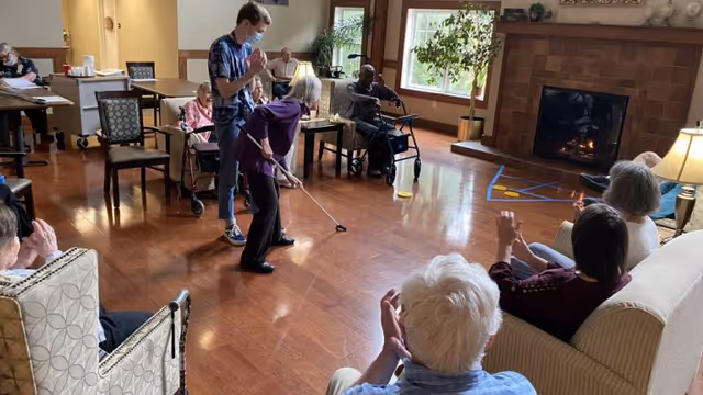 A group of elderly people and a caregiver wearing a mask are gathered in a spacious room with wooden floors and a fireplace. One elderly woman is playing a game with a stick, while others watch and clap. The room has comfortable chairs, tables, and large windows letting in natural light.