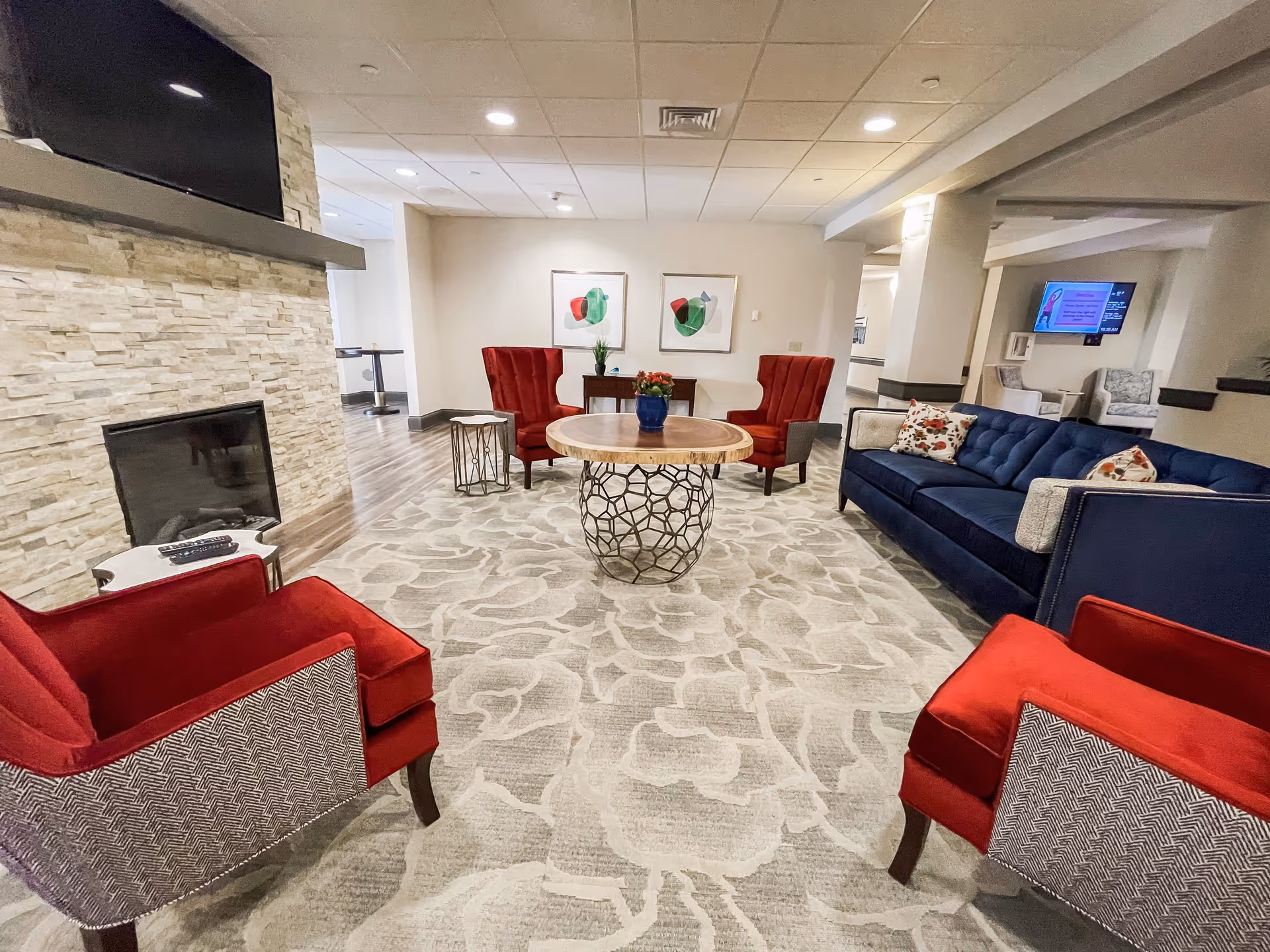 A cozy living room area in The Polaris Community featuring a blue sofa with patterned pillows, four red armchairs, a round wooden coffee table with a decorative metal base, a fireplace with a mounted TV above it, and two abstract paintings on the wall. The room has a light-colored patterned carpet and recessed ceiling lights.
