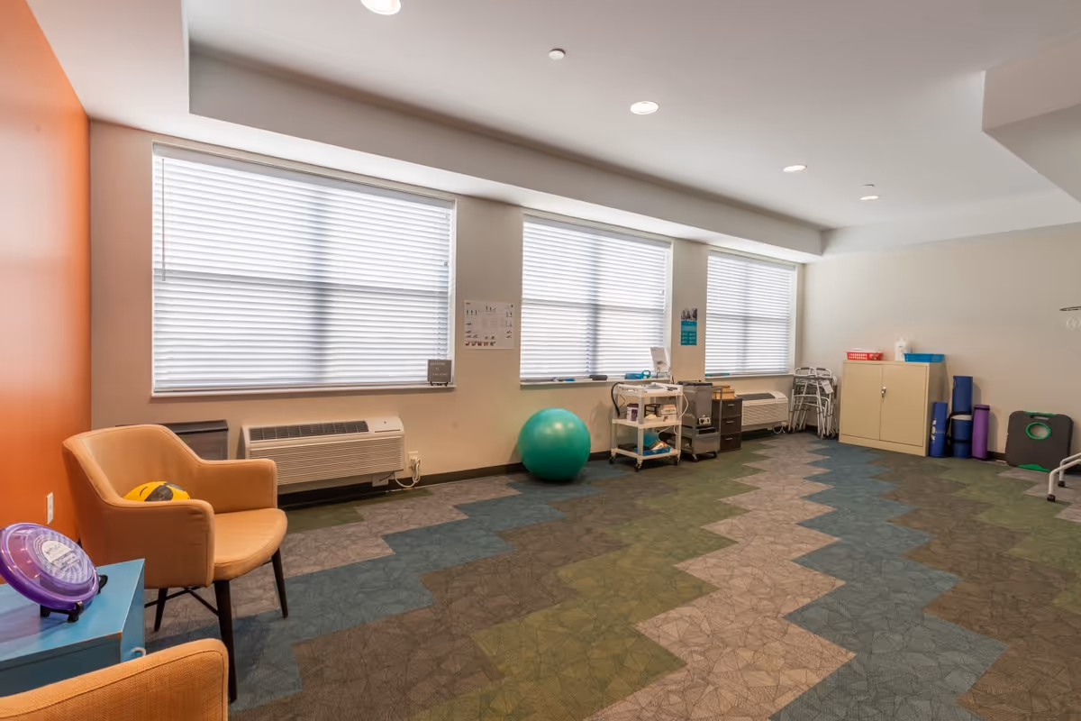 A spacious room with large windows covered by blinds, featuring a patterned carpet floor in shades of green, blue, and gray. The room contains two beige armchairs, a green exercise ball, a small cart with medical or therapy supplies, a cabinet, several rolled-up exercise mats, and walkers. The walls are painted white with one orange accent wall.