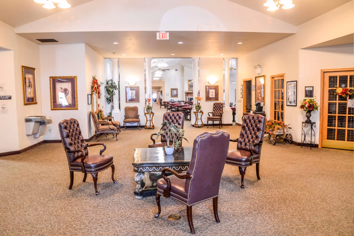 A spacious and well-lit common area with four brown leather tufted chairs arranged around a dark wooden coffee table with a small potted plant on it. The room has beige walls, carpeted floors, framed artwork on the walls, and several other chairs and tables visible in the background. There are decorative plants and floral arrangements placed around the room, and ceiling lights provide warm illumination.