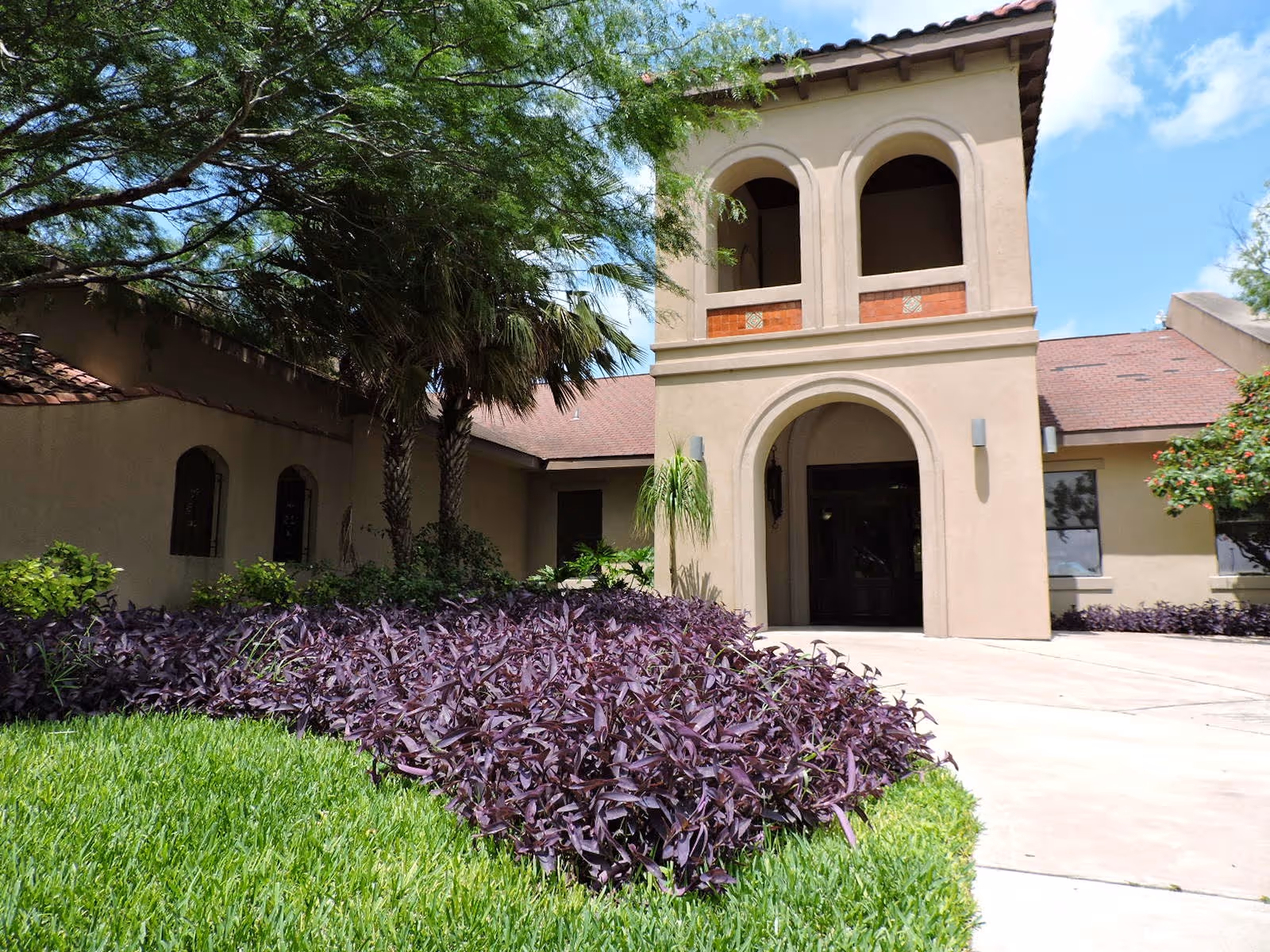 Exterior view of The Bridges at Mission Assisted Living Homes building entrance with arched doorway, beige walls, and a red-tiled roof. The foreground features green grass and purple-leafed plants, with trees providing shade on the left side.