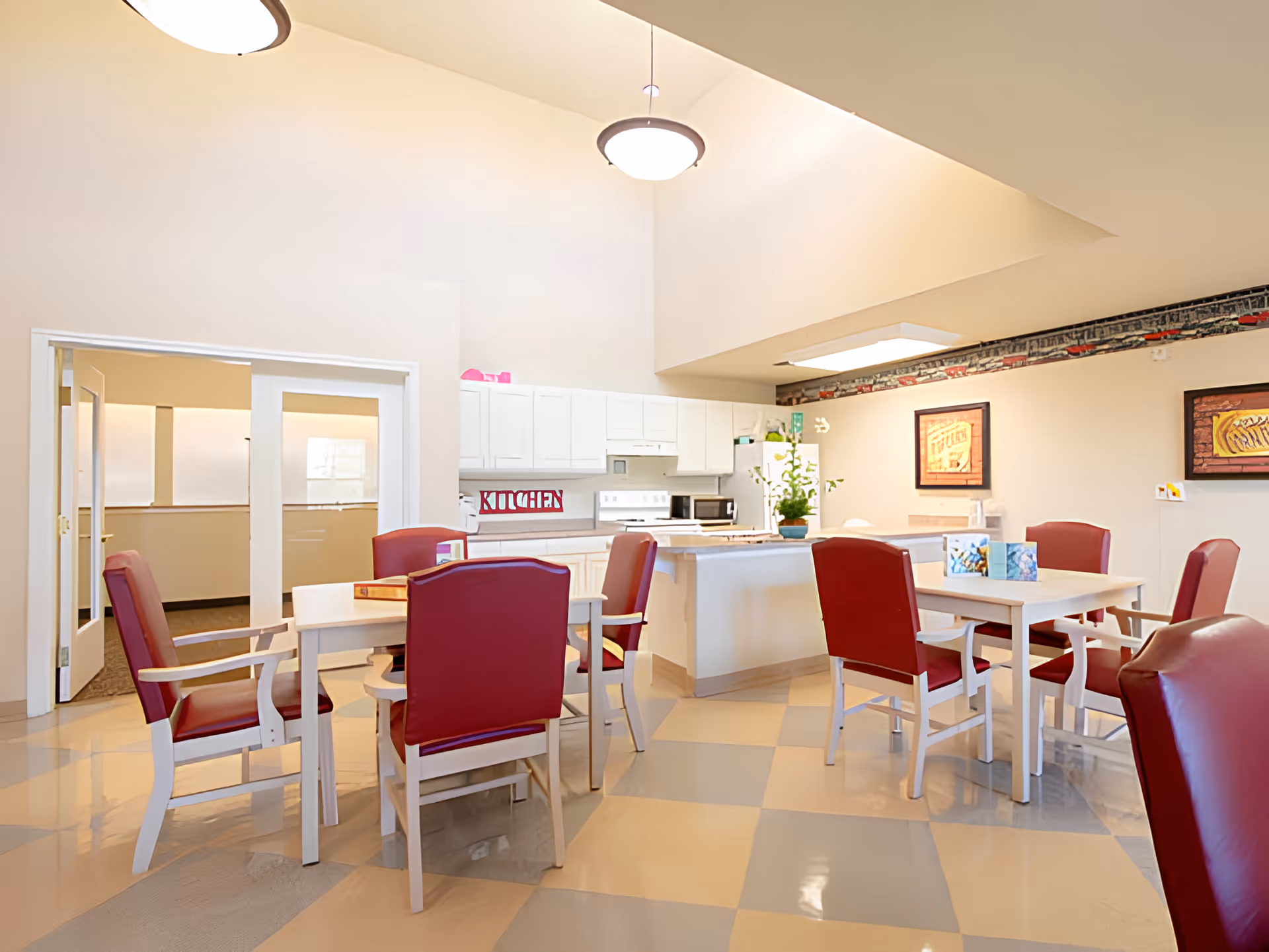 A bright communal kitchen and dining area with white cabinets, a refrigerator, microwave, and stove. There are two white tables surrounded by red cushioned chairs. The floor has a checkered pattern in beige and gray. The walls are decorated with framed artwork and a border featuring a train design near the ceiling.