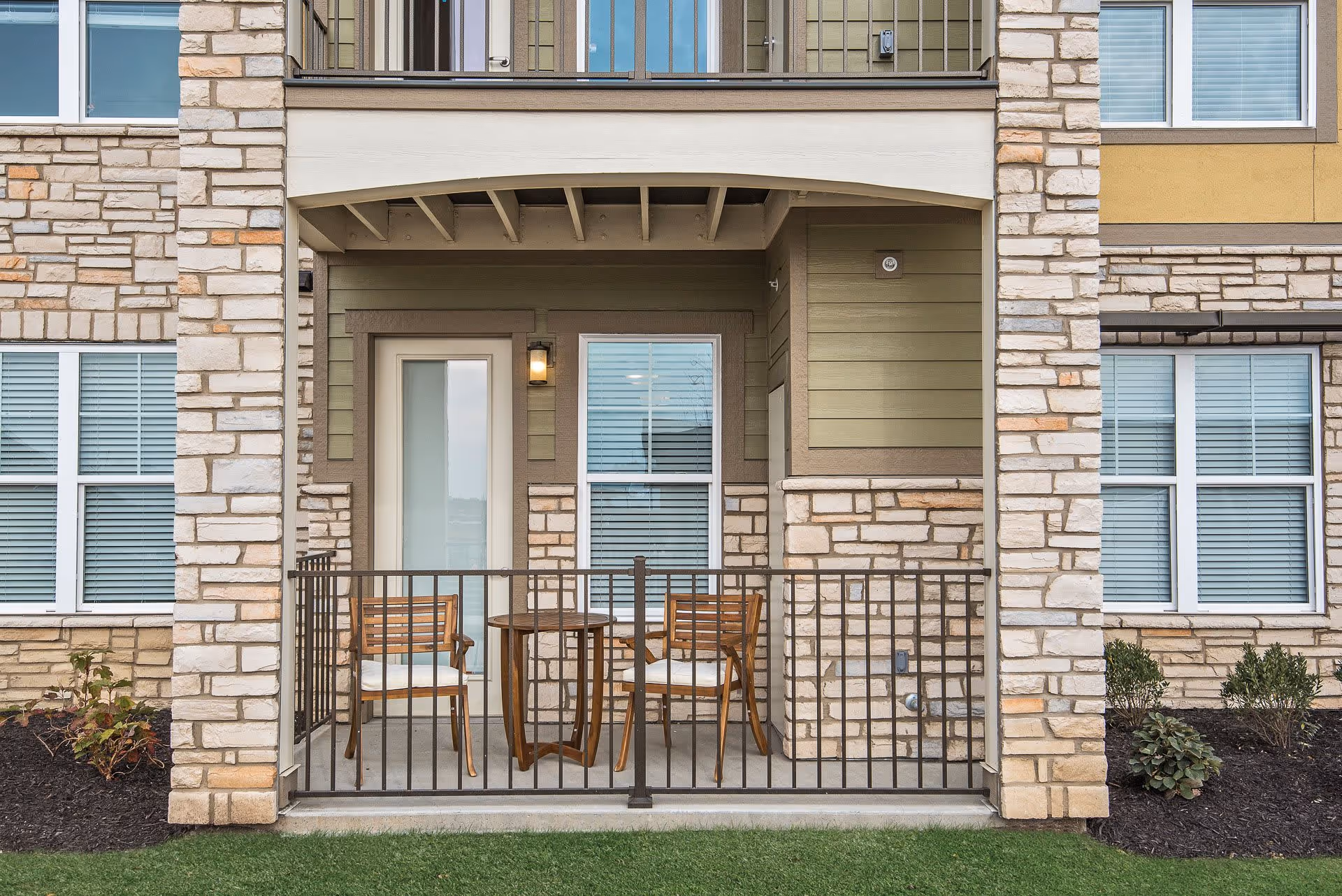 Small outdoor patio area with two wooden chairs and a round wooden table, enclosed by a black metal railing. The patio is part of a building with stone and green siding exterior walls, and there is a door and window behind the patio.