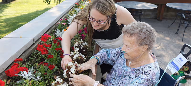 An elderly woman in a wheelchair with an oxygen tube is gardening with the help of a younger woman. They are tending to a flower bed filled with red and white flowers outdoors on a sunny day.