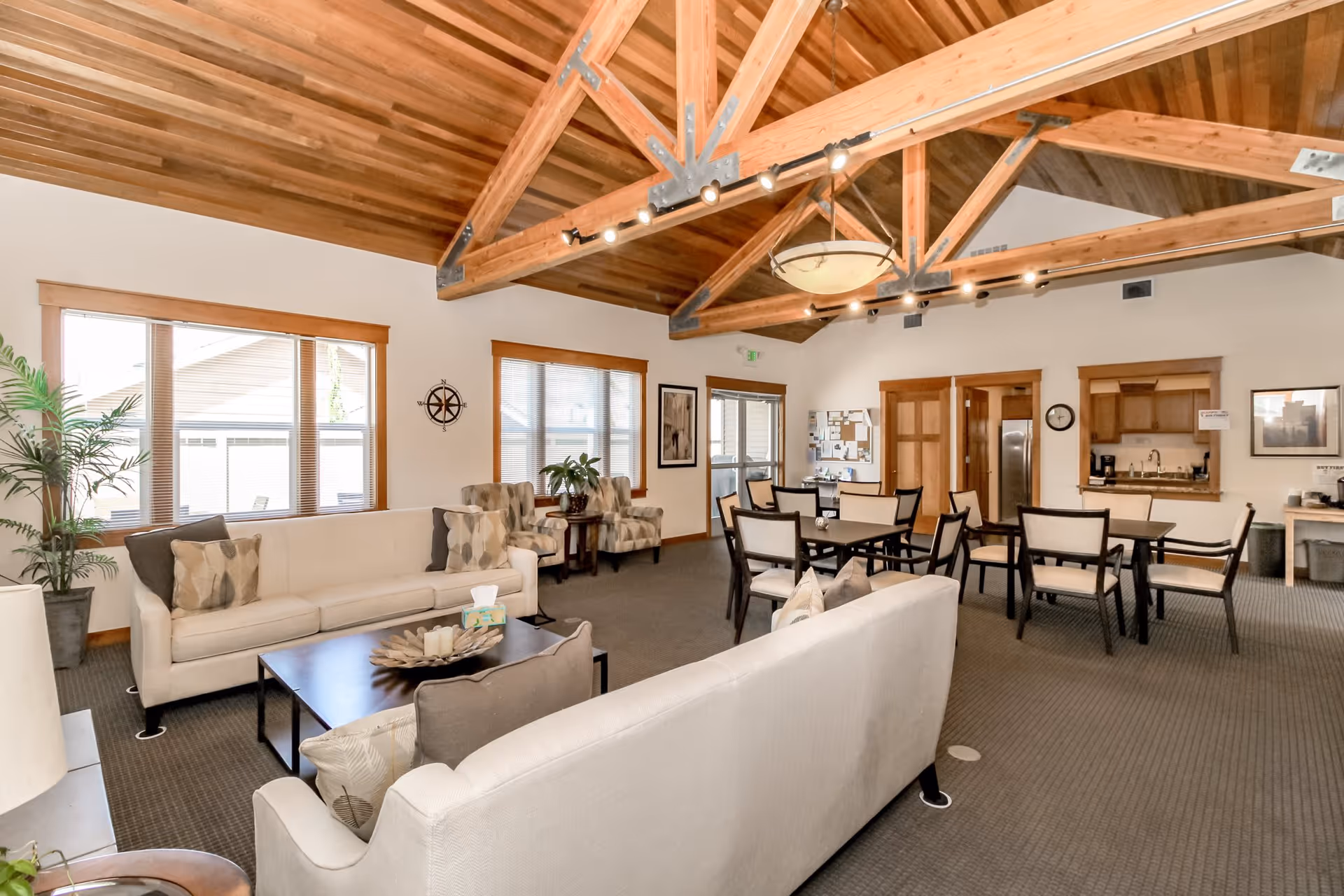 A spacious senior living common area with high wooden vaulted ceilings and exposed beams. The room features two beige sofas with patterned throw pillows, a dark coffee table with decorative items, two patterned armchairs, and a dining area with multiple tables and chairs. Large windows allow natural light to fill the room, and there are plants and framed artwork on the walls. A small kitchen area is visible in the background.