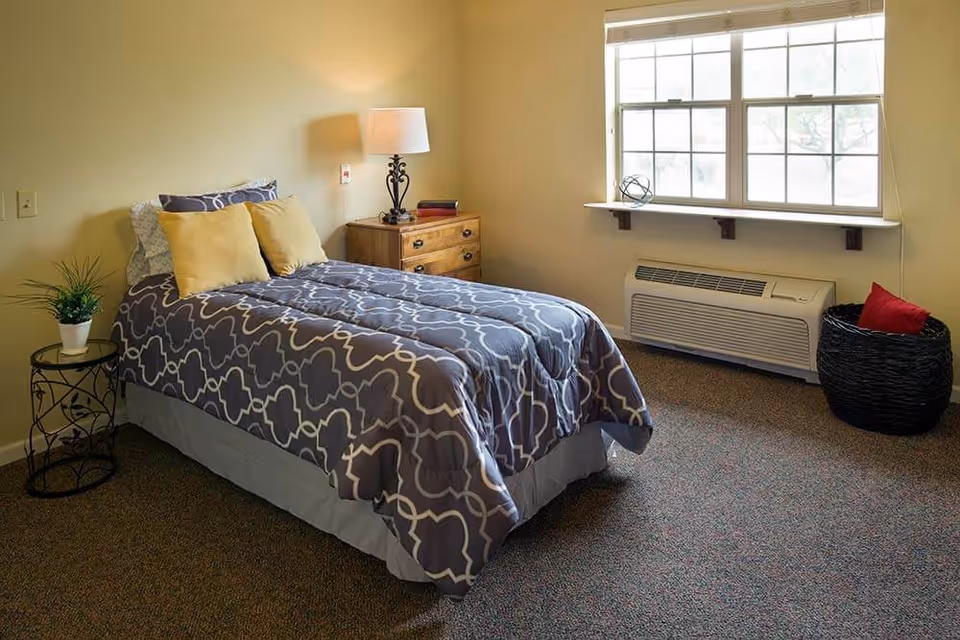 A neatly made single bed with a gray and white patterned comforter and two yellow pillows in a small bedroom. Next to the bed is a wooden nightstand with a decorative lamp and a book. A small round table with a potted plant is beside the bed. A window with blinds lets in natural light, and below the window is a heating/cooling unit. A black woven basket with a red pillow is on the floor near the window.