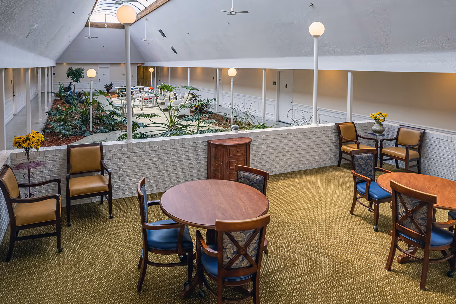 Indoor common area in a senior living facility with round wooden tables and chairs arranged on a carpeted floor. A low white brick wall separates this seating area from a garden-like space with plants and additional seating under a high ceiling with skylights and globe lights on poles.