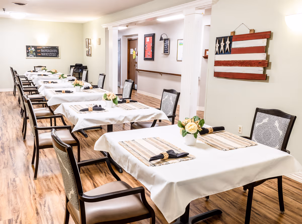 A dining room in a senior living facility with several tables covered in white tablecloths, each set with placemats, napkins, and small flower arrangements. The room has wooden flooring, light-colored walls, and decorative elements including a rustic American flag wall hanging and framed pictures. Chairs with patterned upholstery are arranged around the tables.