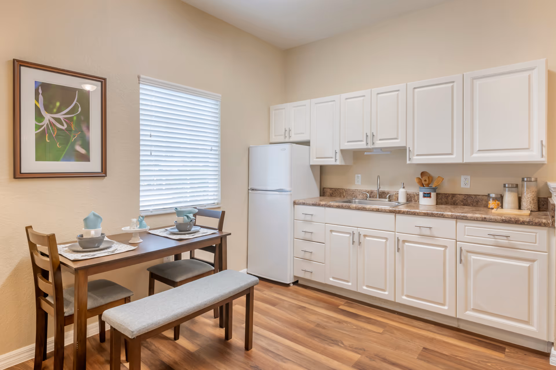 Small kitchen with white cabinets and refrigerator, a dining table set with two chairs and a bench, and framed art on the wall.