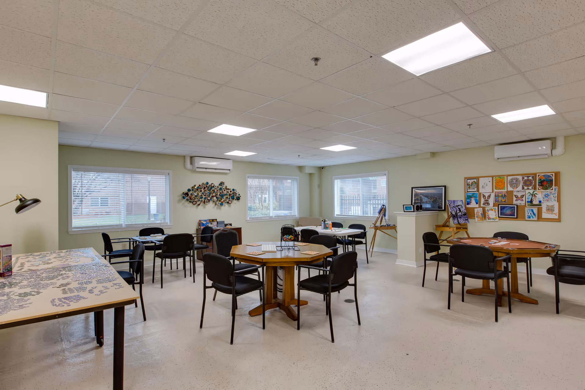 A well-lit activity room with multiple tables and chairs arranged for group activities. One table has a partially completed puzzle, and another has art supplies. The walls feature windows with blinds, a bulletin board with colorful artwork, and decorative wall art. The room has a clean, organized appearance with a light-colored floor and ceiling tiles.