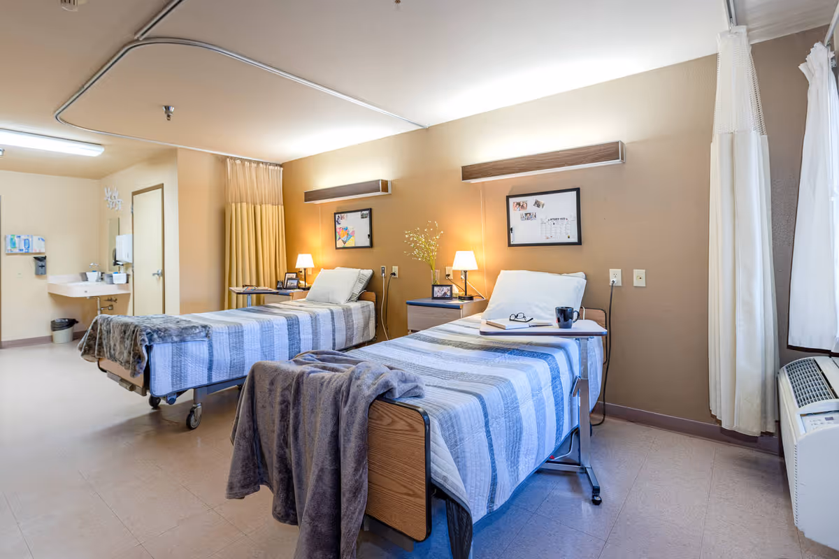 A bright and clean room with two hospital-style beds, each with striped blue and white bedding and a gray blanket. Each bed has a small bedside table with a lamp, framed photos, and other personal items. There is a sink and a door in the background, a curtain divider between the beds, and a window with white curtains on the right side. The room has beige walls and a tiled floor.