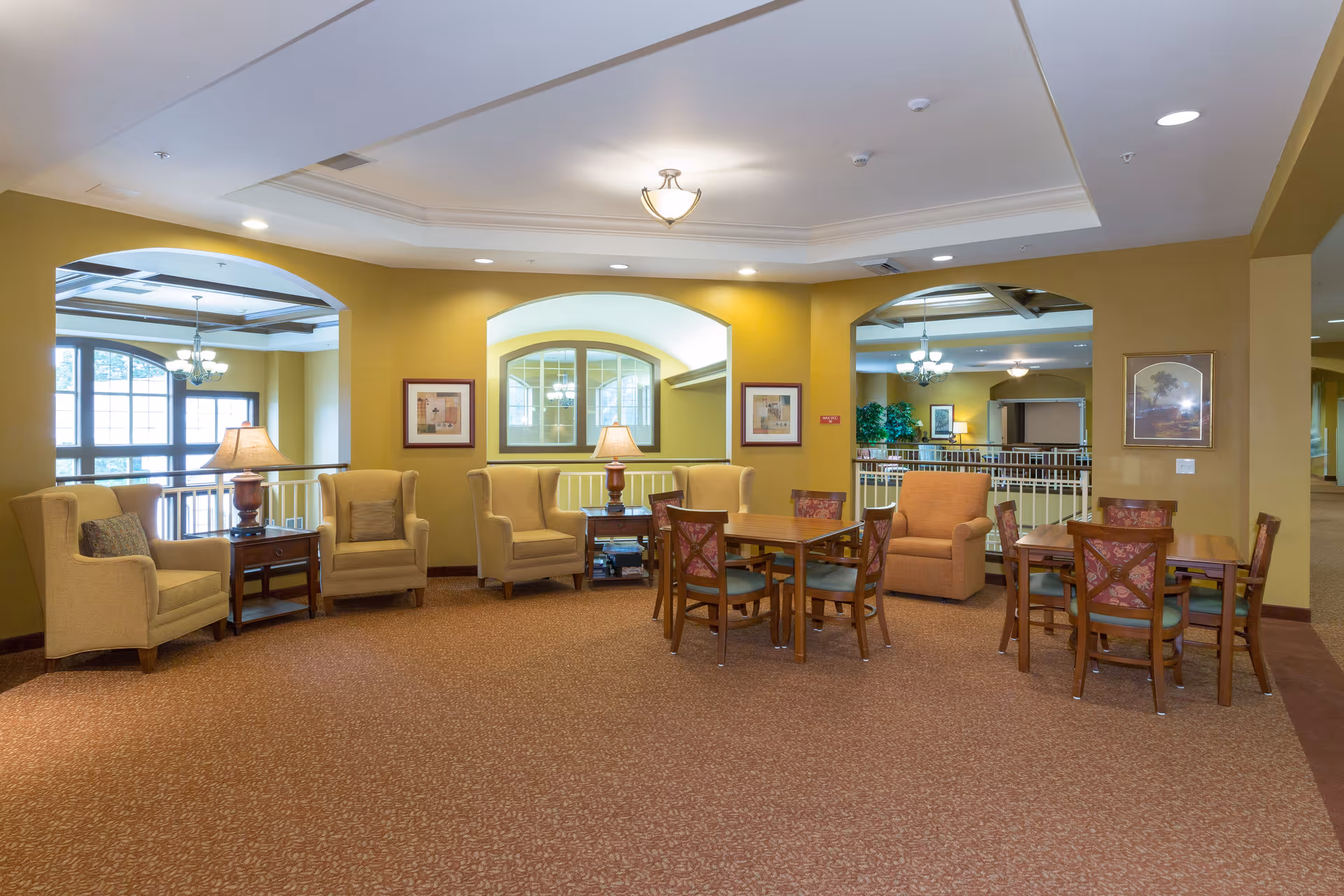 A spacious common area in a senior living facility with beige armchairs and wooden tables with chairs arranged on a patterned carpet. The walls are painted yellow, and there are framed pictures and table lamps on side tables. Large windows and ceiling lights provide ample lighting.