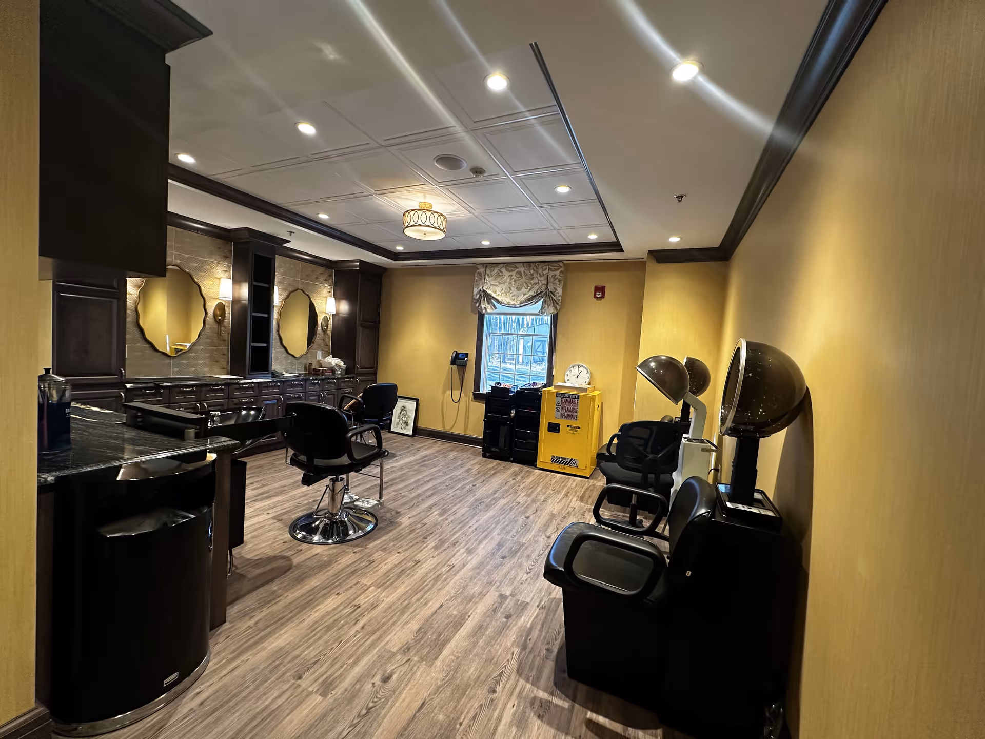 Interior view of a salon room with wooden flooring and yellow walls. The room features salon chairs, hair drying stations with hooded dryers, a counter with mirrors and wall sconces, and a window with a decorative curtain. There is a yellow flammable materials cabinet and a clock near the window.
