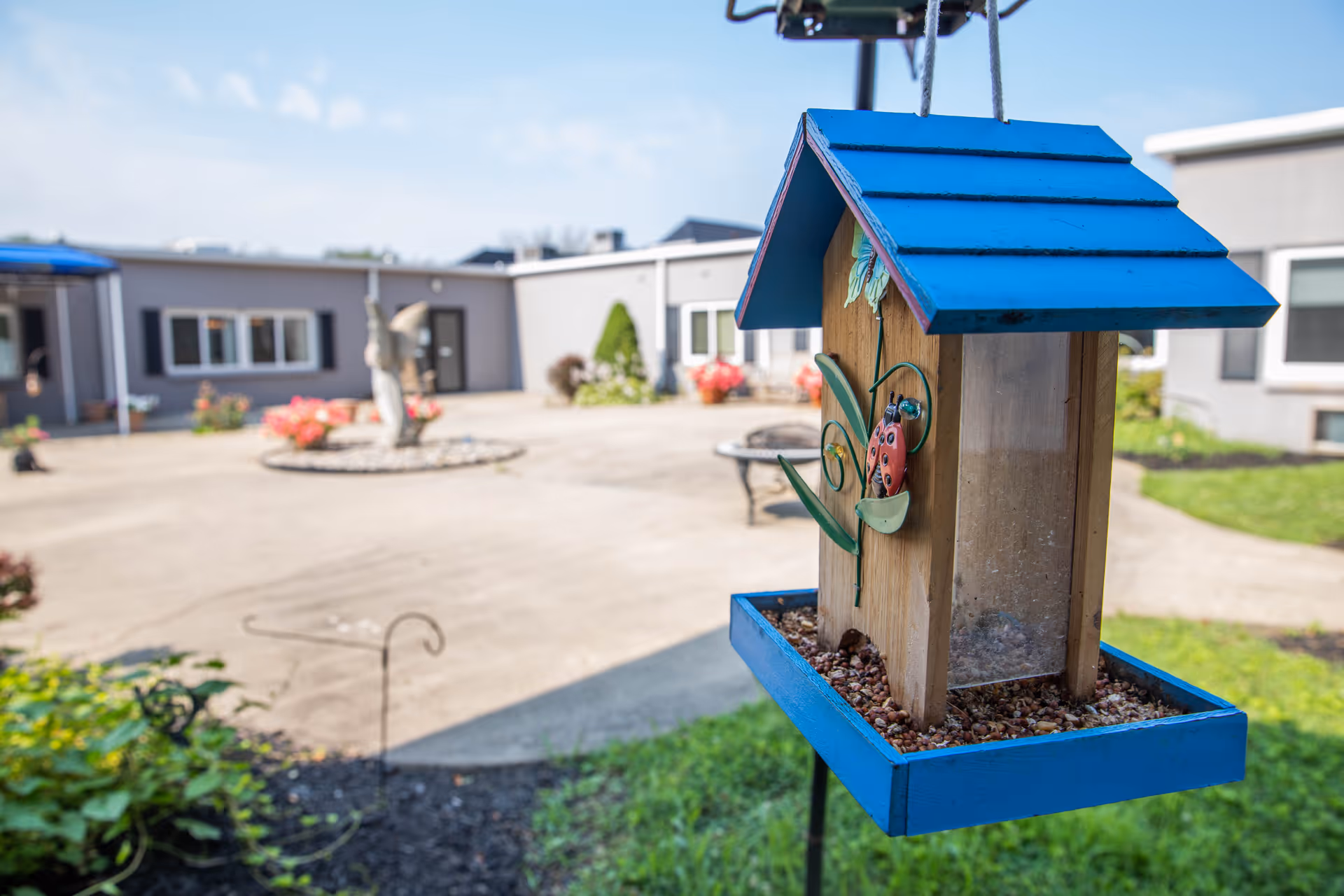 A close-up of a blue bird feeder filled with seeds hanging outdoors with a courtyard and building in the background under a clear sky.