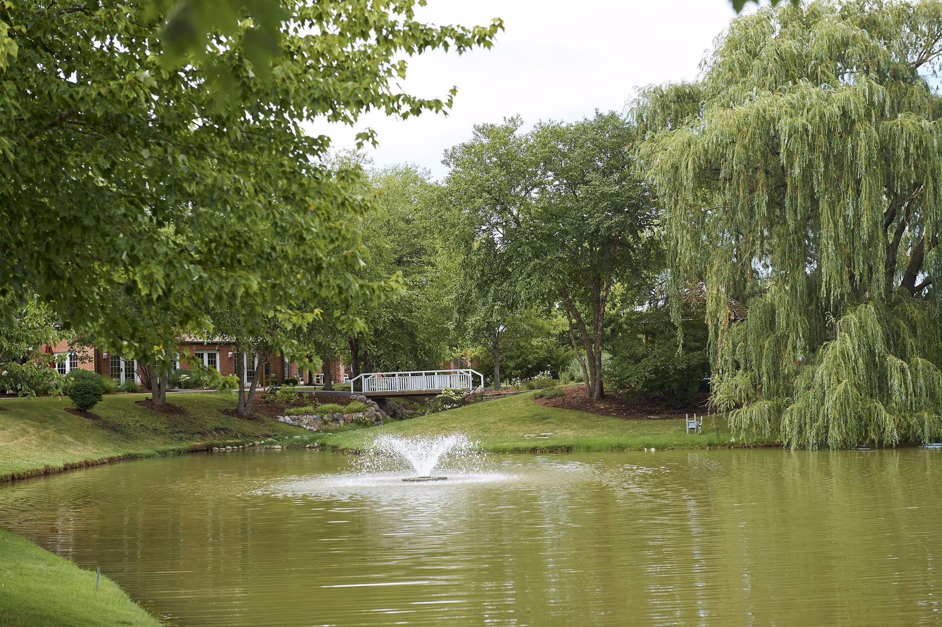 A peaceful outdoor scene at Peace Village featuring a pond with a water fountain in the center, surrounded by lush green trees and grass. A white footbridge crosses a small stream in the background, with a large weeping willow tree on the right side.