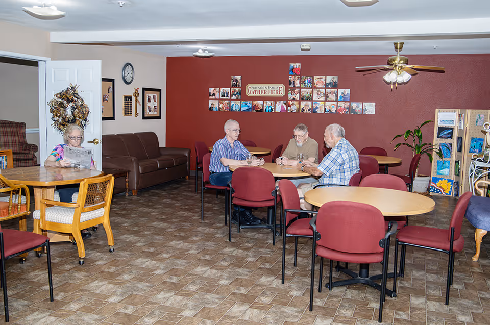 A common area in a retirement community with several round tables and red chairs. Three elderly men are sitting at one table playing cards, while an elderly woman is sitting alone at another table reading a newspaper. The walls are decorated with framed photos and a sign that reads 'Friends & Family Gather Here.' There is a ceiling fan and a plant in the corner.