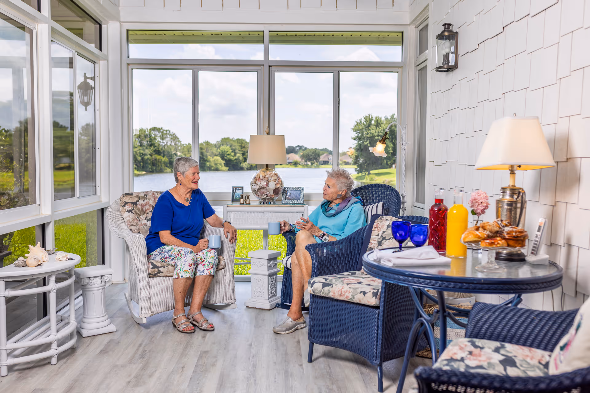Two elderly women sitting and chatting in a bright sunroom with large windows overlooking a lake and greenery. The room is furnished with wicker chairs with floral cushions, a small table with drinks and pastries, and decorative lamps. The atmosphere is cozy and inviting.