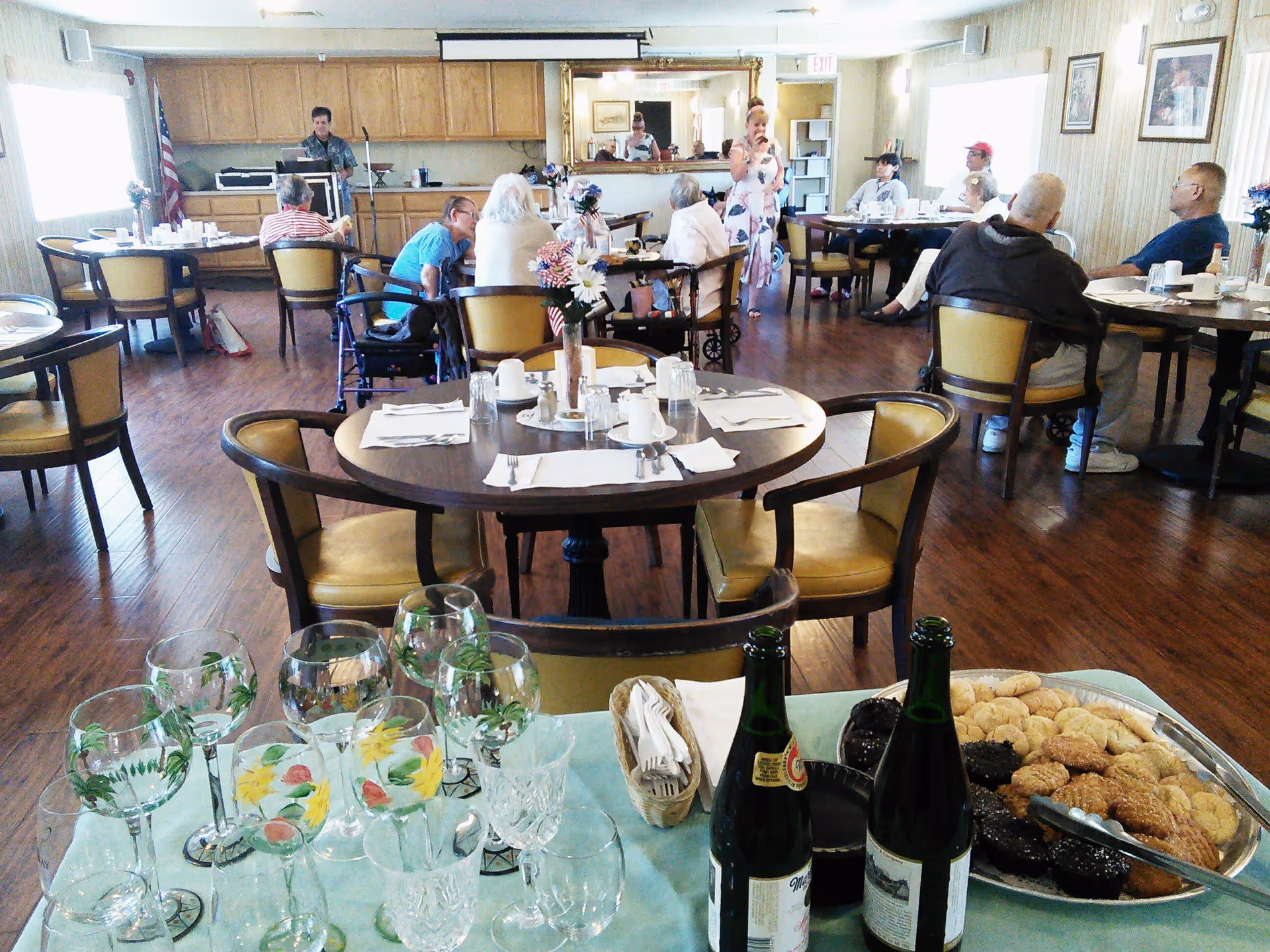 Residents and staff in a senior living dining room with set tables and a refreshment station in the foreground.