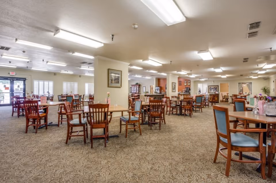 A spacious dining room with multiple wooden tables and chairs arranged neatly. The room is well-lit with fluorescent ceiling lights and has carpeted flooring with a patterned design. There are windows with blinds on one side and a door leading outside. The walls are decorated with framed pictures and there is a counter area in the background.